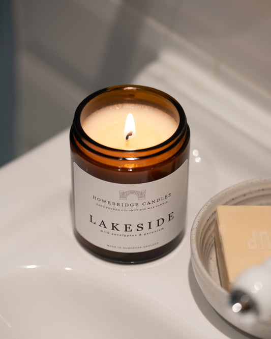 Lit candle in a glass jar labeled 'Lakeside' by Howebridge Canldes, on a white bathroom surface.