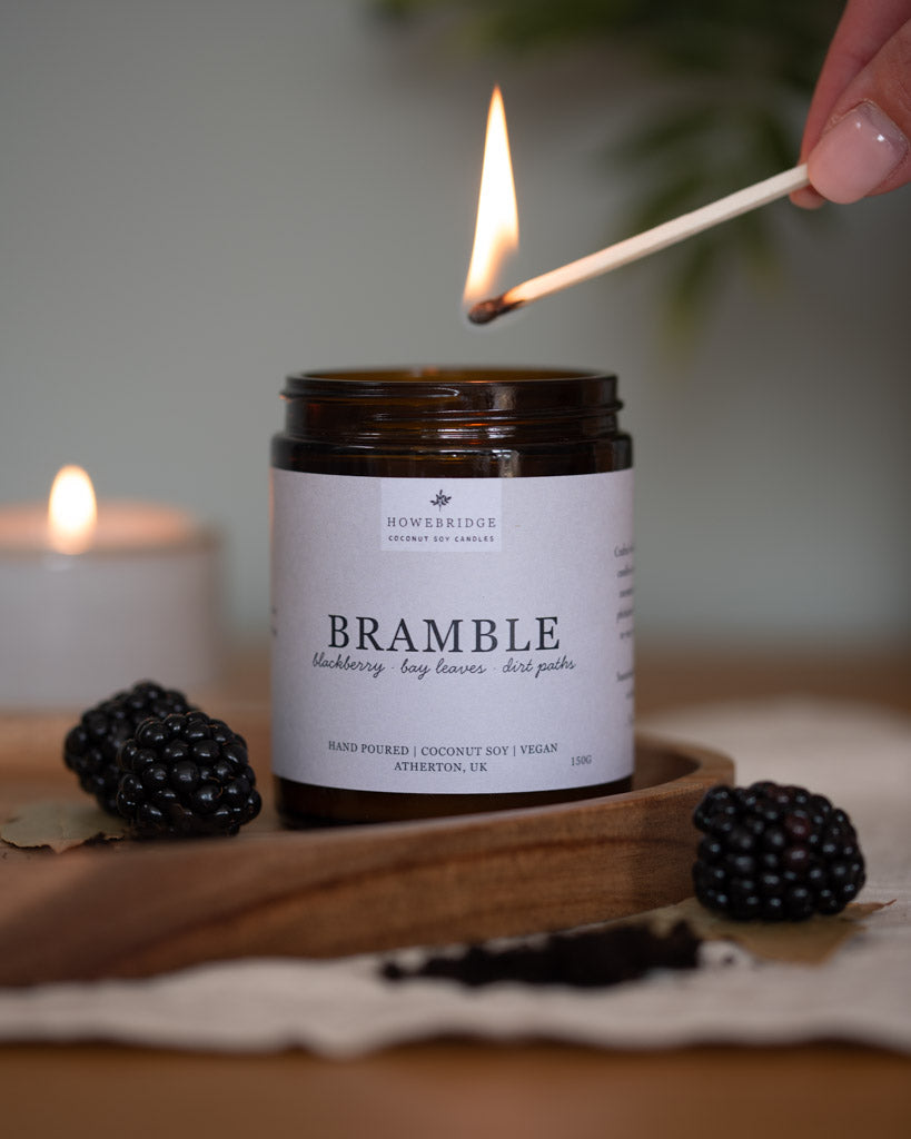 Candle jar labeled 'Bramble' being lit with a match, surrounded by blackberries on a wooden board. Scent: Blackberries, bay leaves and dirt paths. Amber jar with natural white label. Feel like your in a forest in the late summer evening blackberry picking.