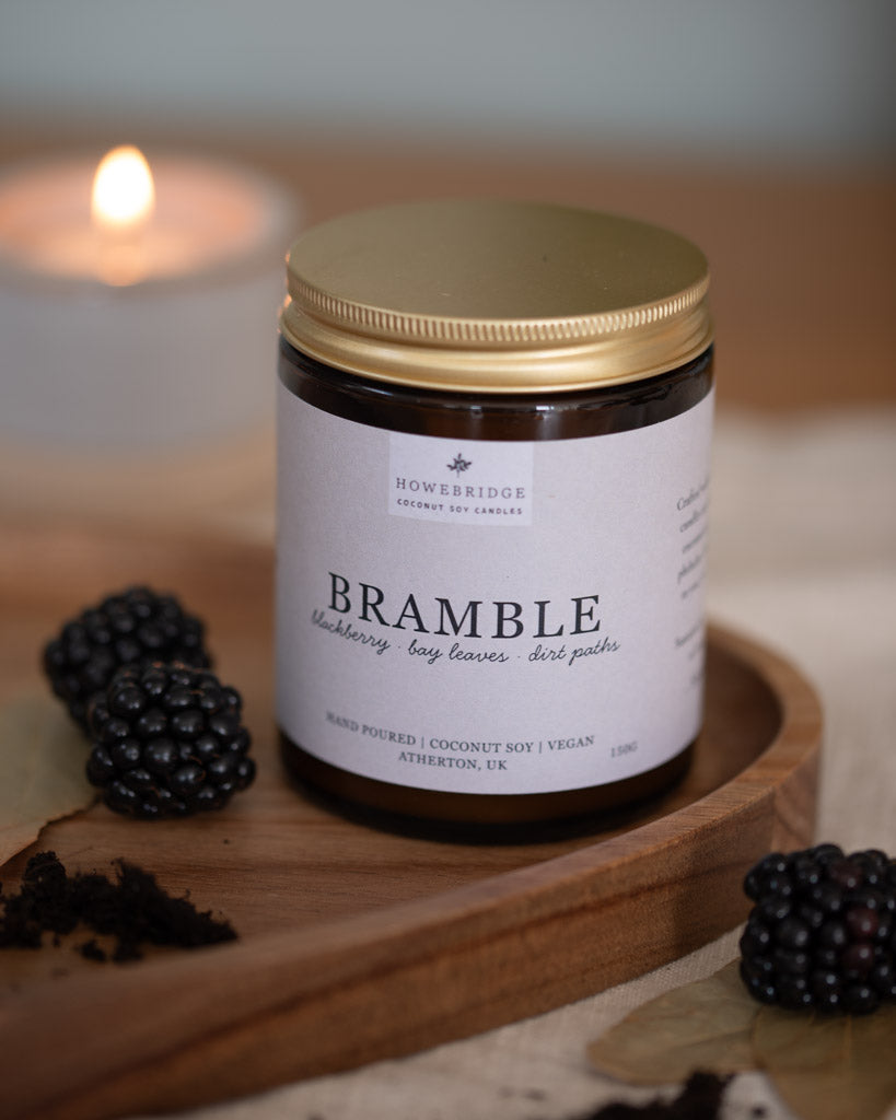 Jar of Bramble candle with gold lid on a wooden board with blackberries, soft focus background. Scent: Blackberries, bay leaves and dirt paths. A sweet, earthy, cosy candle, perfect for summer or autumn. Smells like your blackberry picking in a forest.