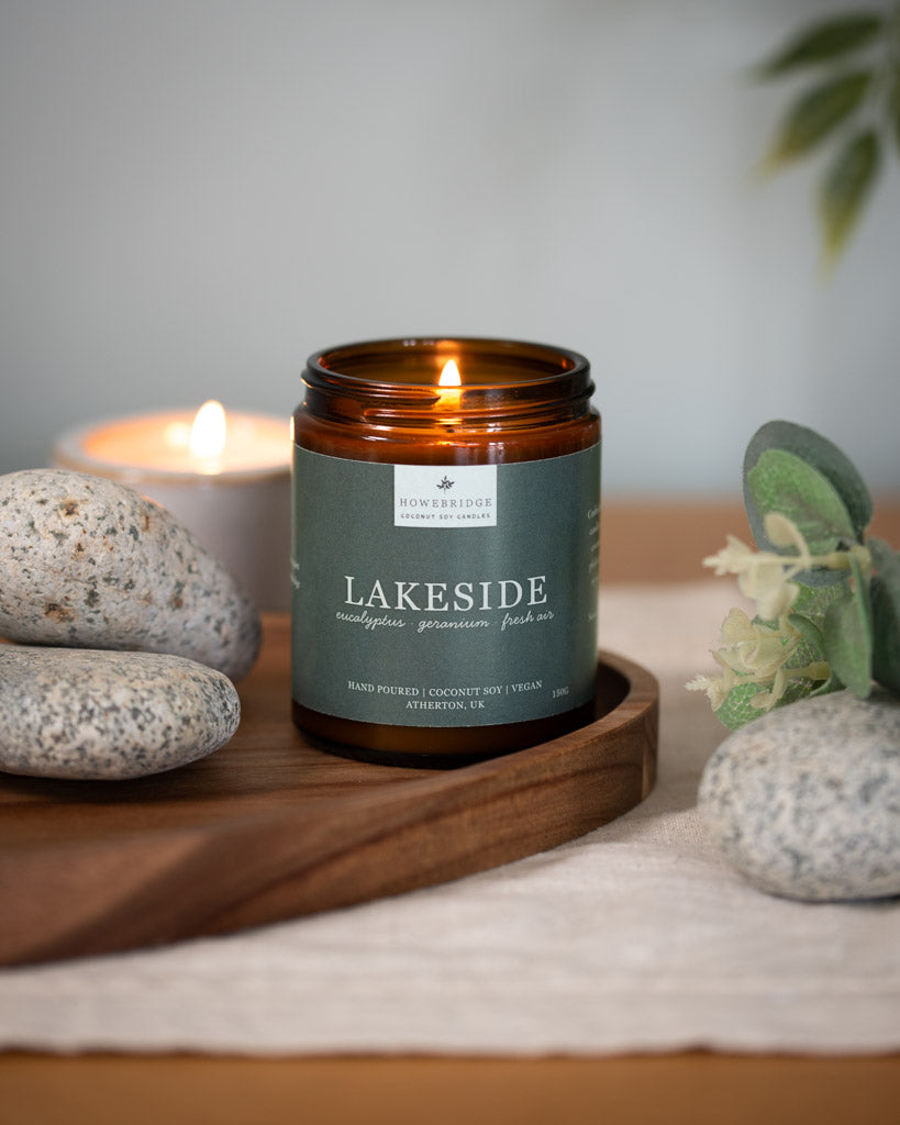 Lakeside candle with the lid off and lit. Surrounded by stones and eucalyptus. Scent: eucalyptus, geranium and fresh air. Smells like sitting by a waterfall or a lake. Bringing a little bit of the outdoors into your home.