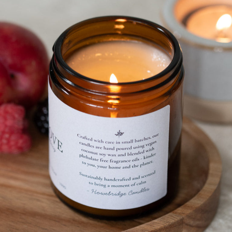 Rhubarb wild grove candle lit on a wooden board surrounded by summer fruits. Amber jar with a natural wrap around label. Label on the side talks about how the candle is hand poured, coconut soy wax and phthalate free.