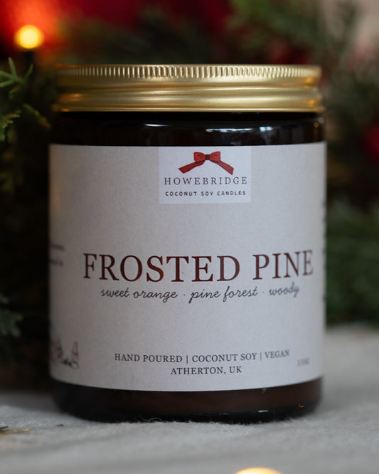 Christmas candle called Frosted Pine. Hand poured by Howebridge Candles, in a glass amber jar and gold lid with a festive background. Sustainably made, vegan friendly and toxin-free.