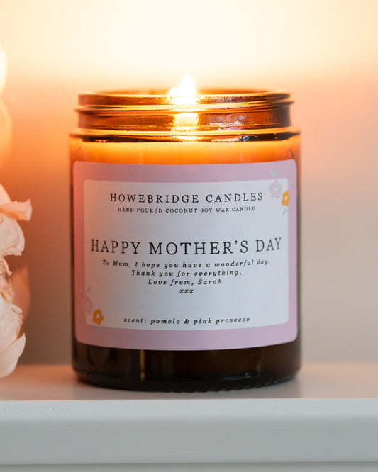 Personalised mother's day candle, made by Howebridge Candles. A glass amber jar candle and label saying happy mothers day, lit and sat on a white shelf with pink flowers.