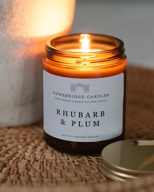 rhubarb candle in a glass amber jar, the candle is lit and sat on a rattan tray.