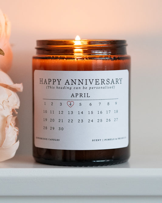 A personalised anniversary candle by Howebridge Candles. A glass amber jar and white label that features a happy anniversary heading and calendar. Lit and sat on a white shelf with pink flowers.