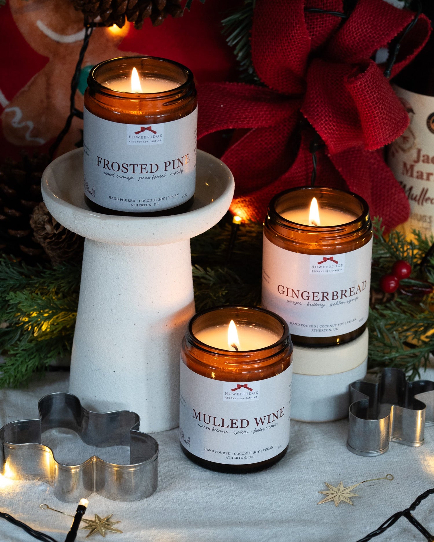 A trio of Howebridge christmas Candles. Mulled wine, Gingerbread and Frosted Pine. All lit in glass amber jars with a festive, decorative background.
