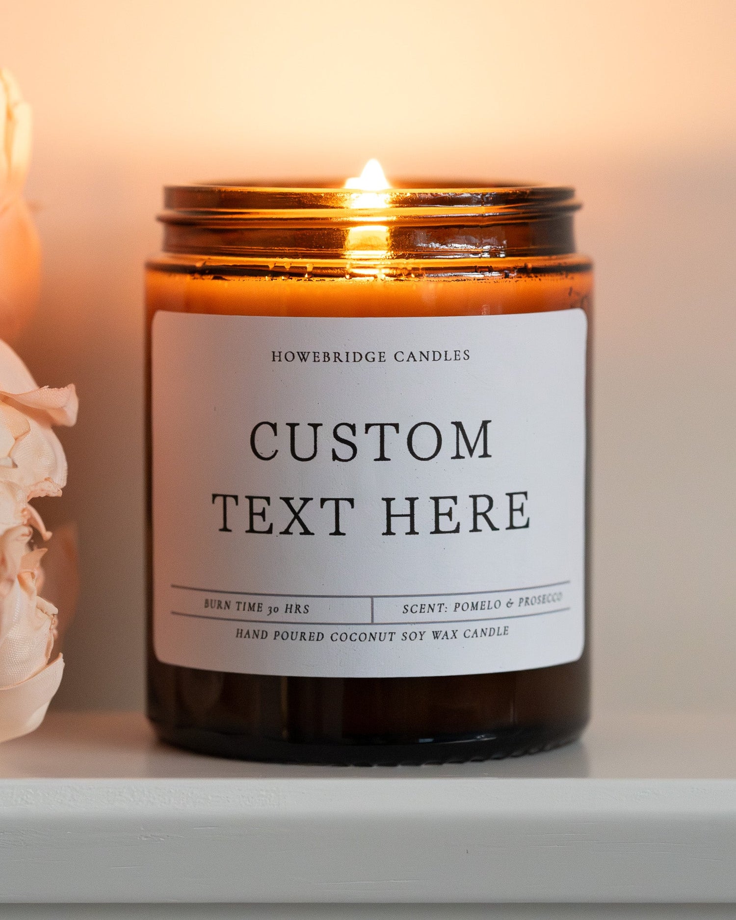 A custom text coconut soy candles by Howebridge Candles. A lit, glass amber jar and white label with black writing, sat on a white shelf with pink flowers.