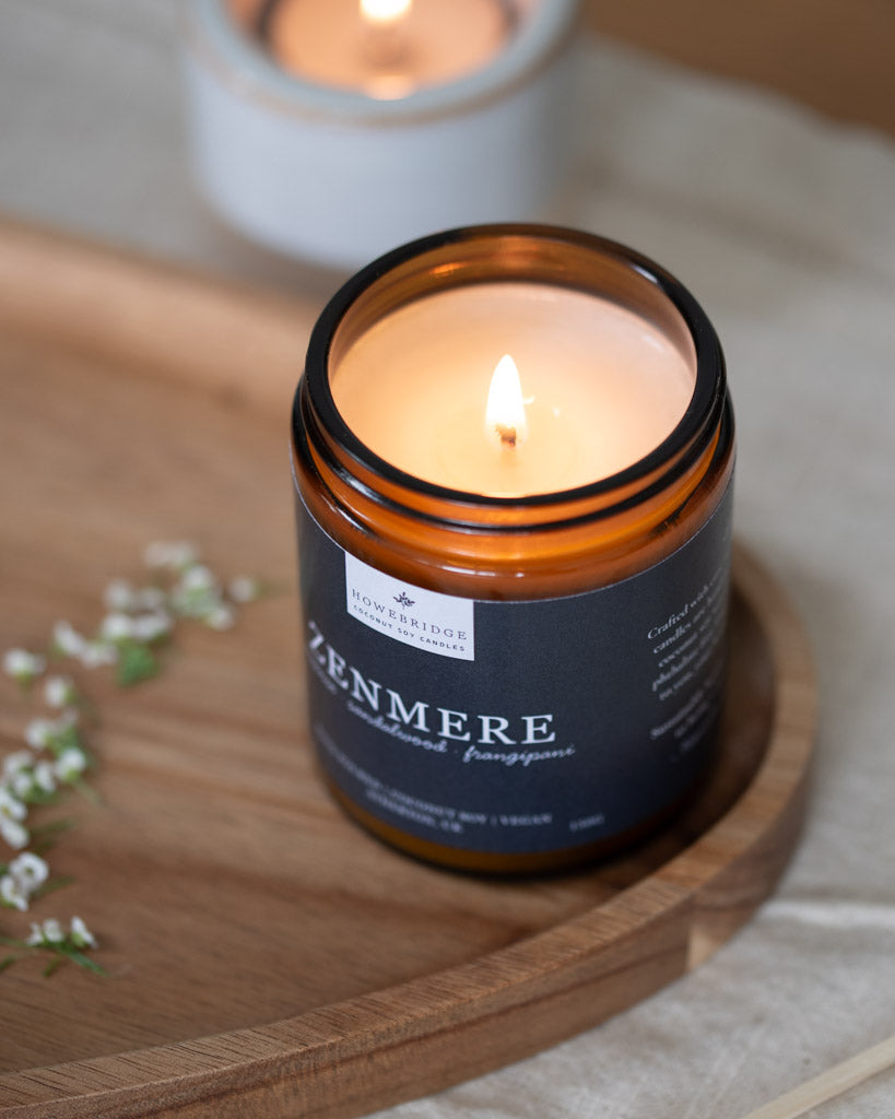 Candle in an amber jar labeled 'Zenmere' on a wooden tray with a soft focus background. Scent is incense, sandalwood and frangipani. A alternative to the traditional incense sticks. The perfect candle to find inner peace and balance and meditate to.