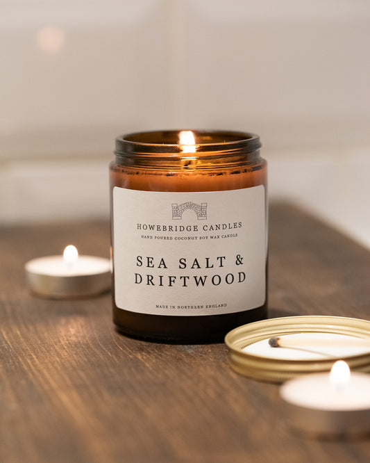 Amber jar candle labeled 'Sea Salt & Driftwood' from Howebridge Candles on a wooden surface with other candles.