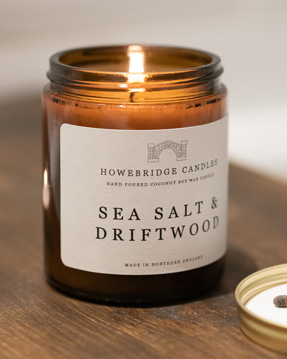 Amber jar candle labeled 'Sea Salt & Driftwood' by Howebridge Candles on a wooden surface.