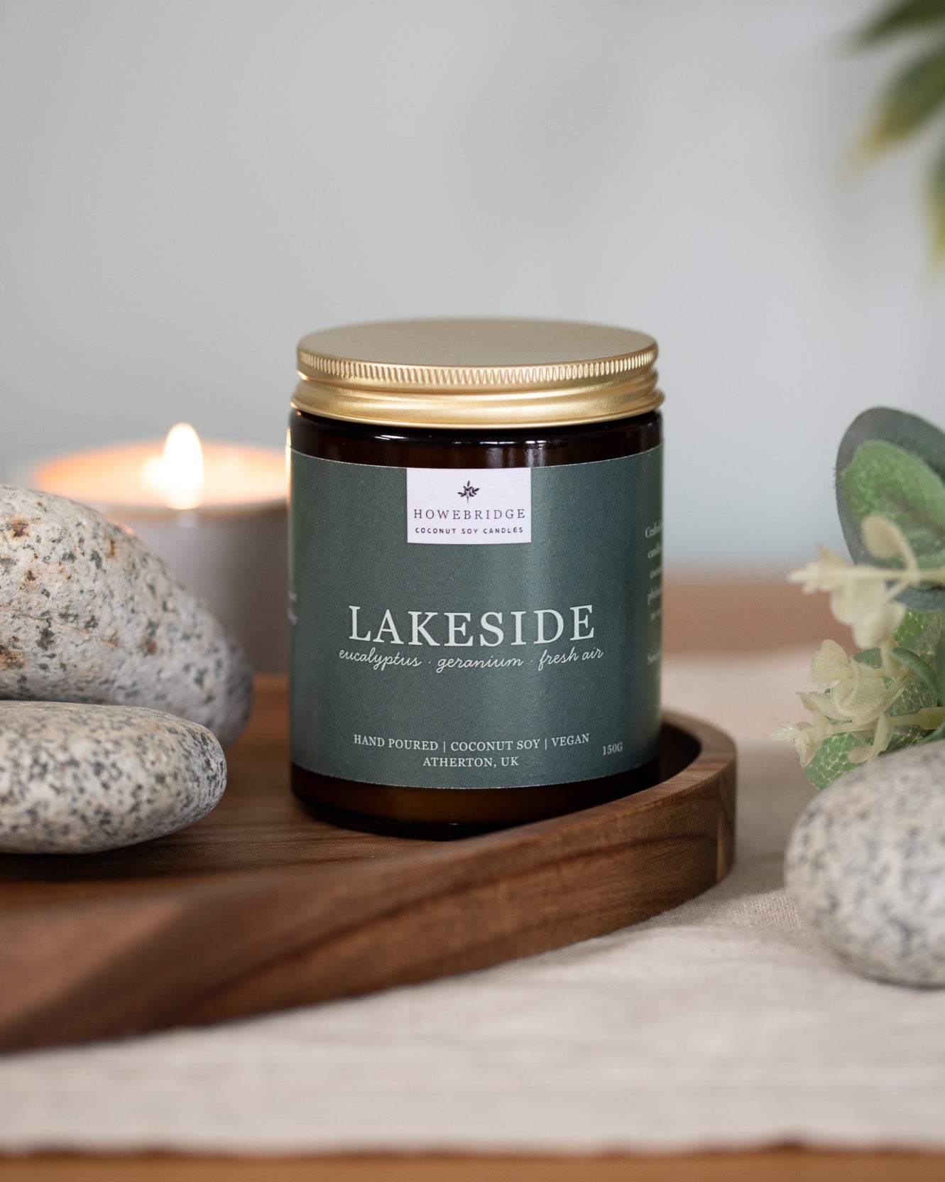 Candle labeled 'Lakeside' on a wooden tray with rocks and eucalyptus. Scent: Eucalyptus, geranium and fresh air. Amber jar with gold lid and a sage green wrap around label. Hand poured, coconut soy wax and vegan.
