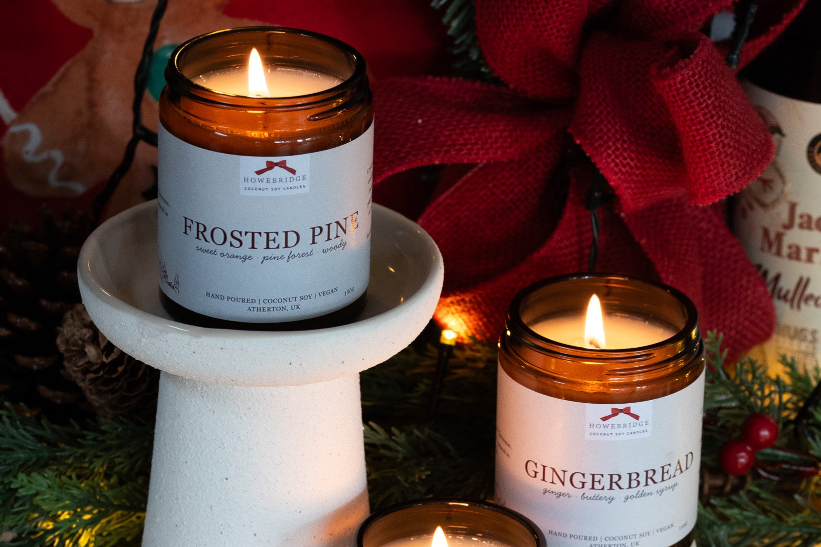 Two lit candles with labels 'Frosted Pine' and 'Gingerbread' on a decorative background. Hand poured into glass amber jars using sustainable coconut soy wax. 