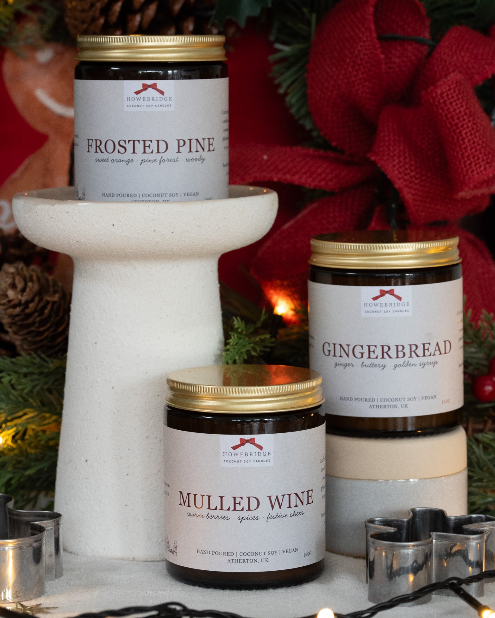 Three holiday-themed candles on a decorative stand with Christmas decorations in the background. Howebridge candles are hand poured into glass amber jars with gold lids, wrapped in a festive label featuring a red bow, fine line festive illustration and scent name.