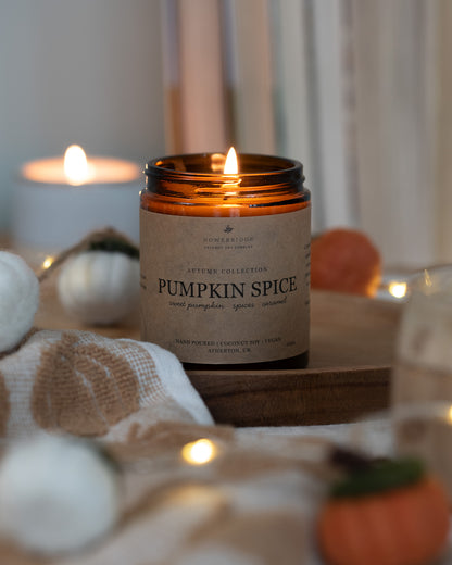 Lit Pumpkin Spice sustainable candle. Kraft label in a glass amber jar. Hand poured coconut soy wax and toxin free fragrances. Cosy and autumnal vibes.