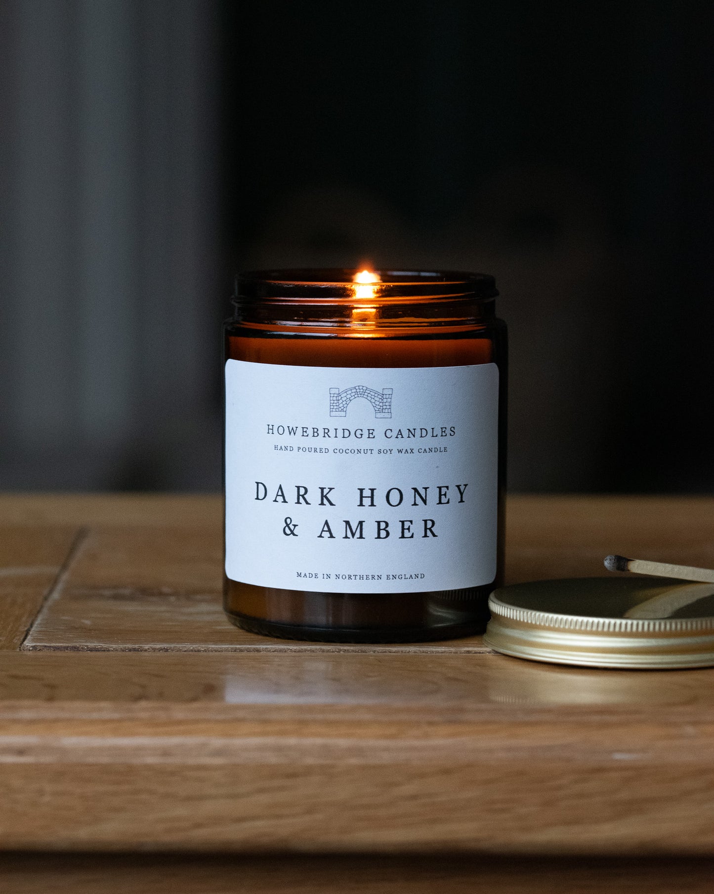 'Dark honey & amber' candle by Howebridge Candles. A glass amber jar and gold lid with white label, sat on a wooden table.