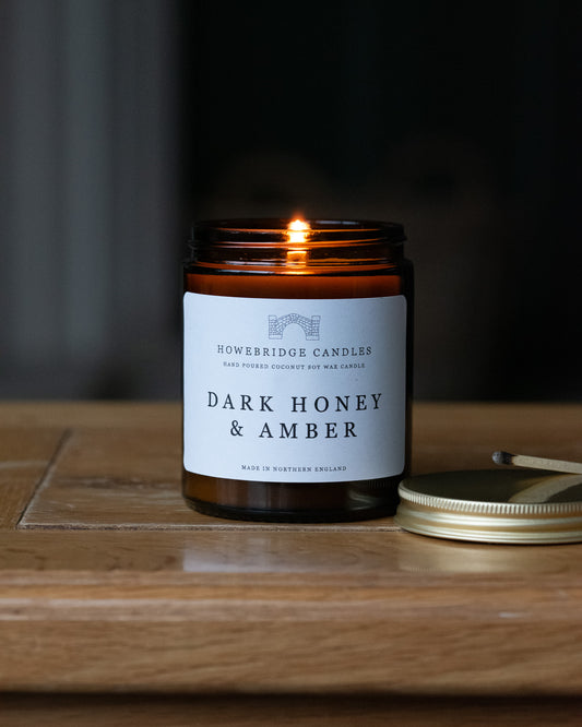 'Dark honey & amber' candle by Howebridge Candles. A glass amber jar and gold lid with white label, sat on a wooden table.