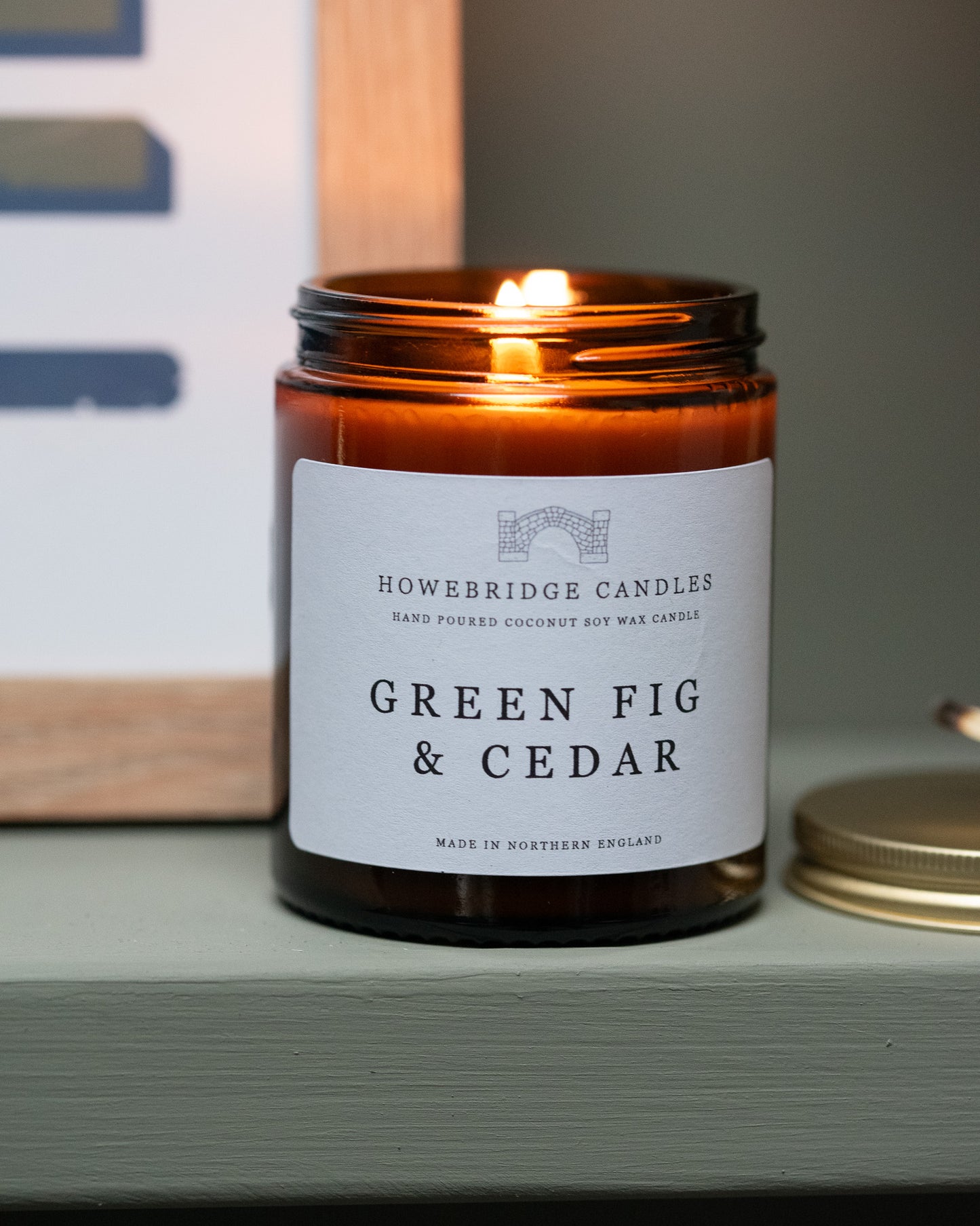 'Green fig & Cedar' candle by Howebridge Candles, in a glass amber jar and gold lid. Sat on a green shelf.