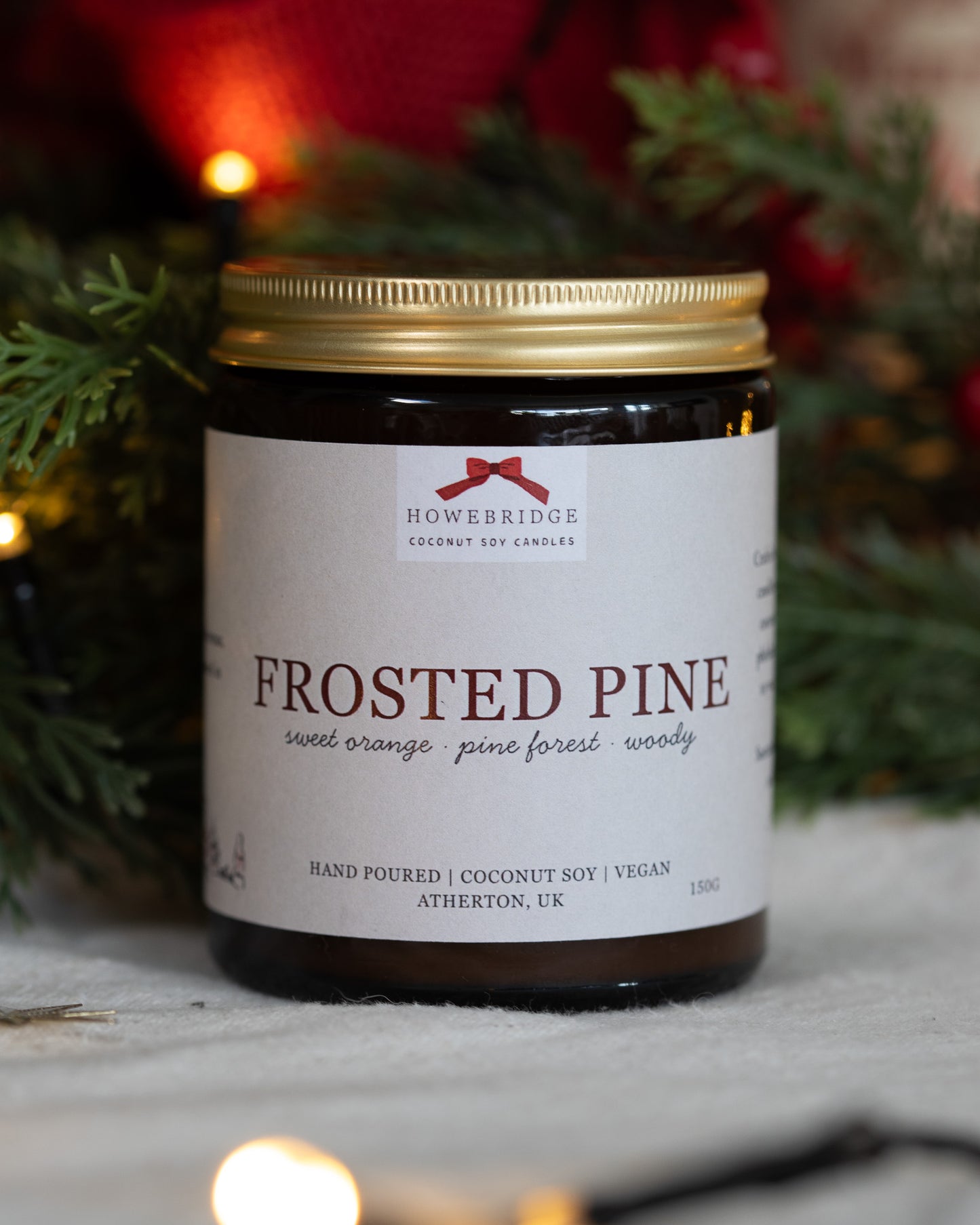 A Christmas candle labeled 'Frosted Pine' by Homebridge Candles with a festive background. Hand poured in a glass amber jar and gold lid. Scent of sweet orange, pine with a woody base.