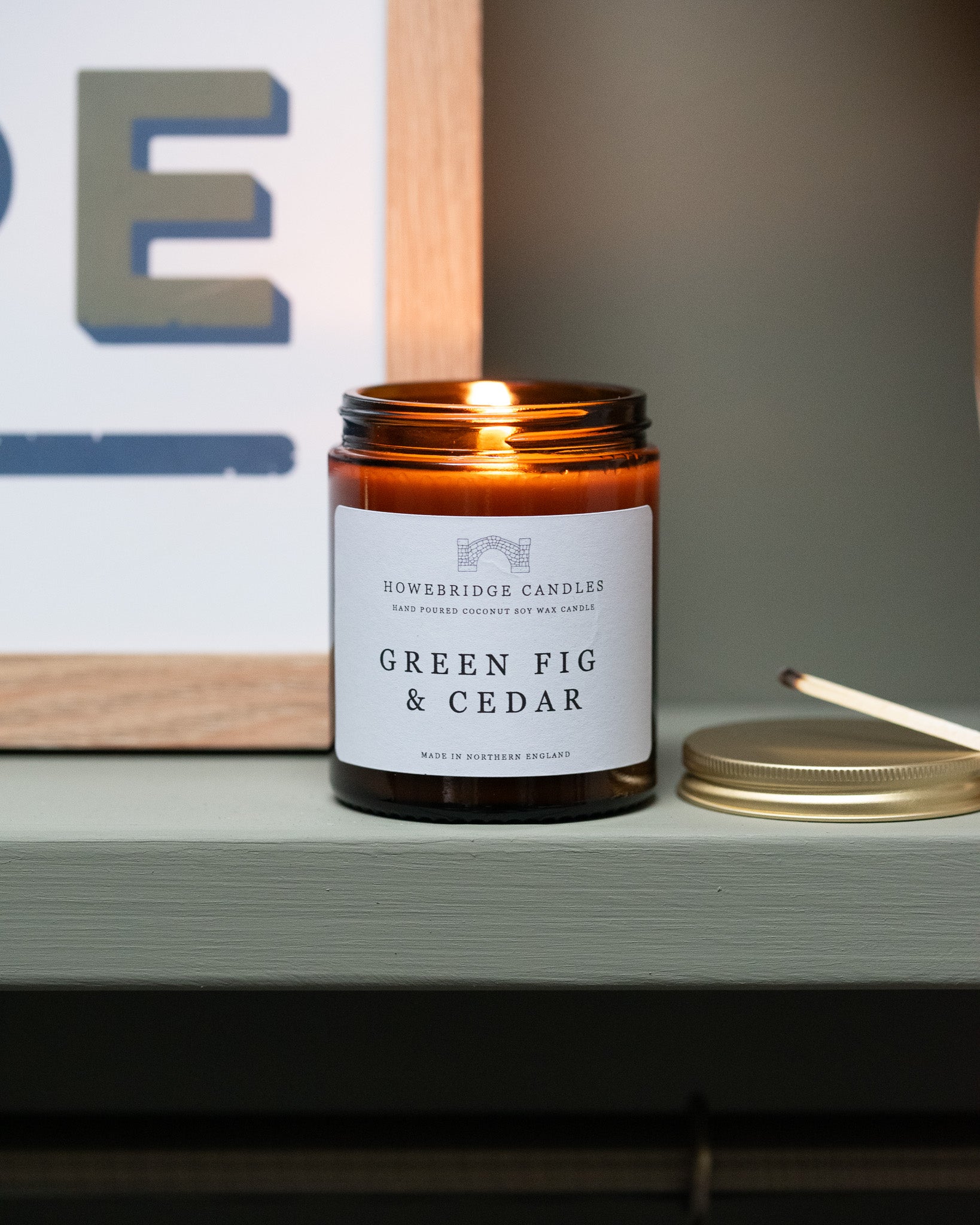 Candle in an amber jar labeled 'Green Fig & Cedar' on a shelf. By Howebridge Candles.