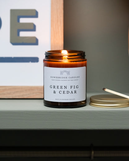 Candle in an amber jar labeled 'Green Fig & Cedar' on a shelf. By Howebridge Candles.