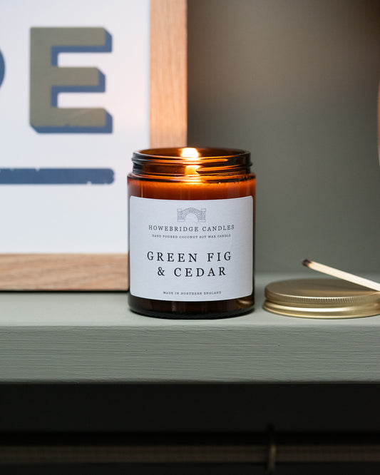 Candle in an amber jar labeled 'Green Fig & Cedar' on a shelf. By Howebridge Candles.