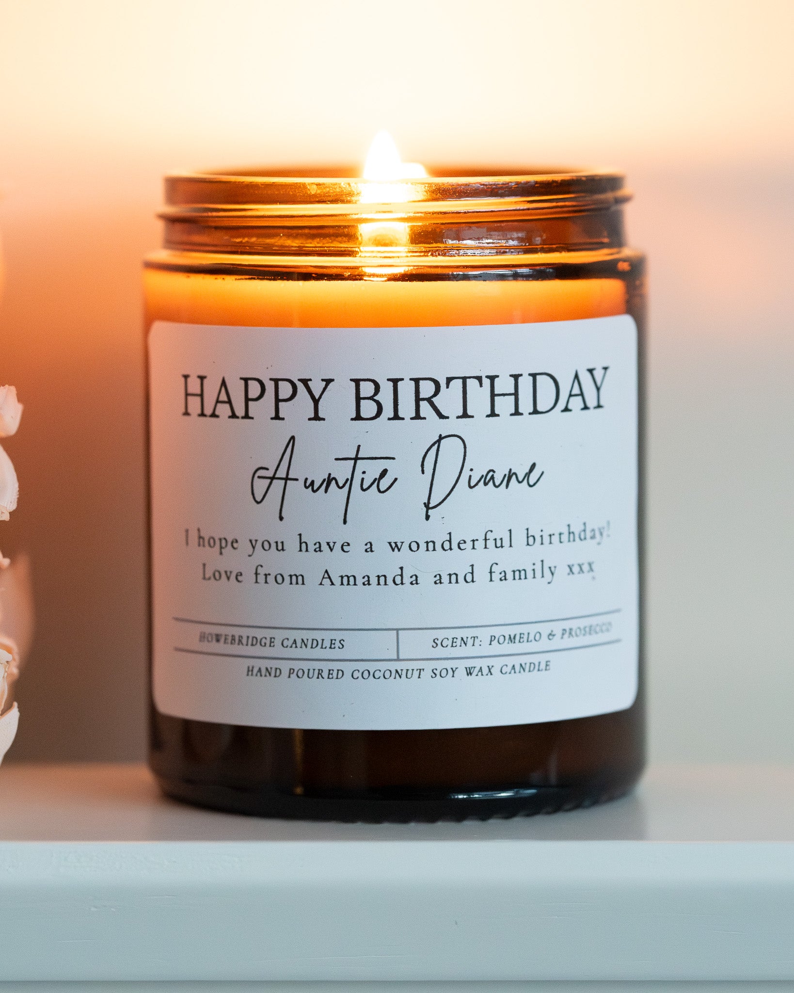 'Happy Birthday' Candle with name and message. Made by Howebridge Candles in a glass amber jar, it's lit and sat on a white shelf with pink flowers.