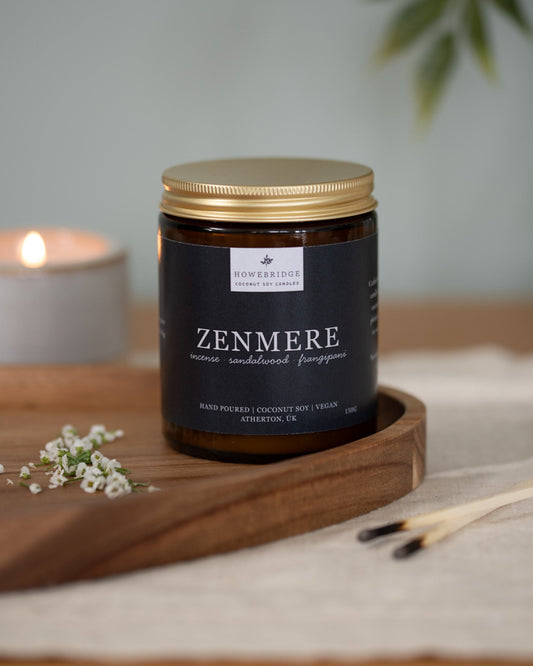 Amber candle jar labeled 'Zenmere' on a wooden tray with a lit candle in the background. Scent is incense, sandalwood and frangipani. A rich Incense and slightly floral, the perfect candle to find inner balance, meditate or light during a yoga session.