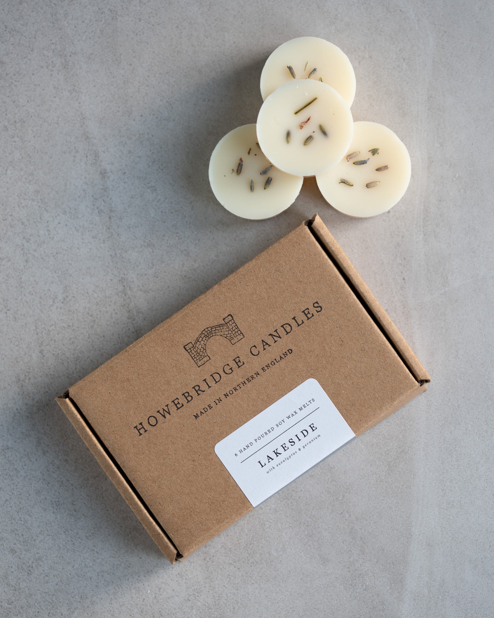 Howebridge Candles Lakeside soy wax melts, stacked and decorated with dried lavender. Next to them is the wax melt packaging, a kraft box stamped with their logo and sealed with a scent descriptive label.