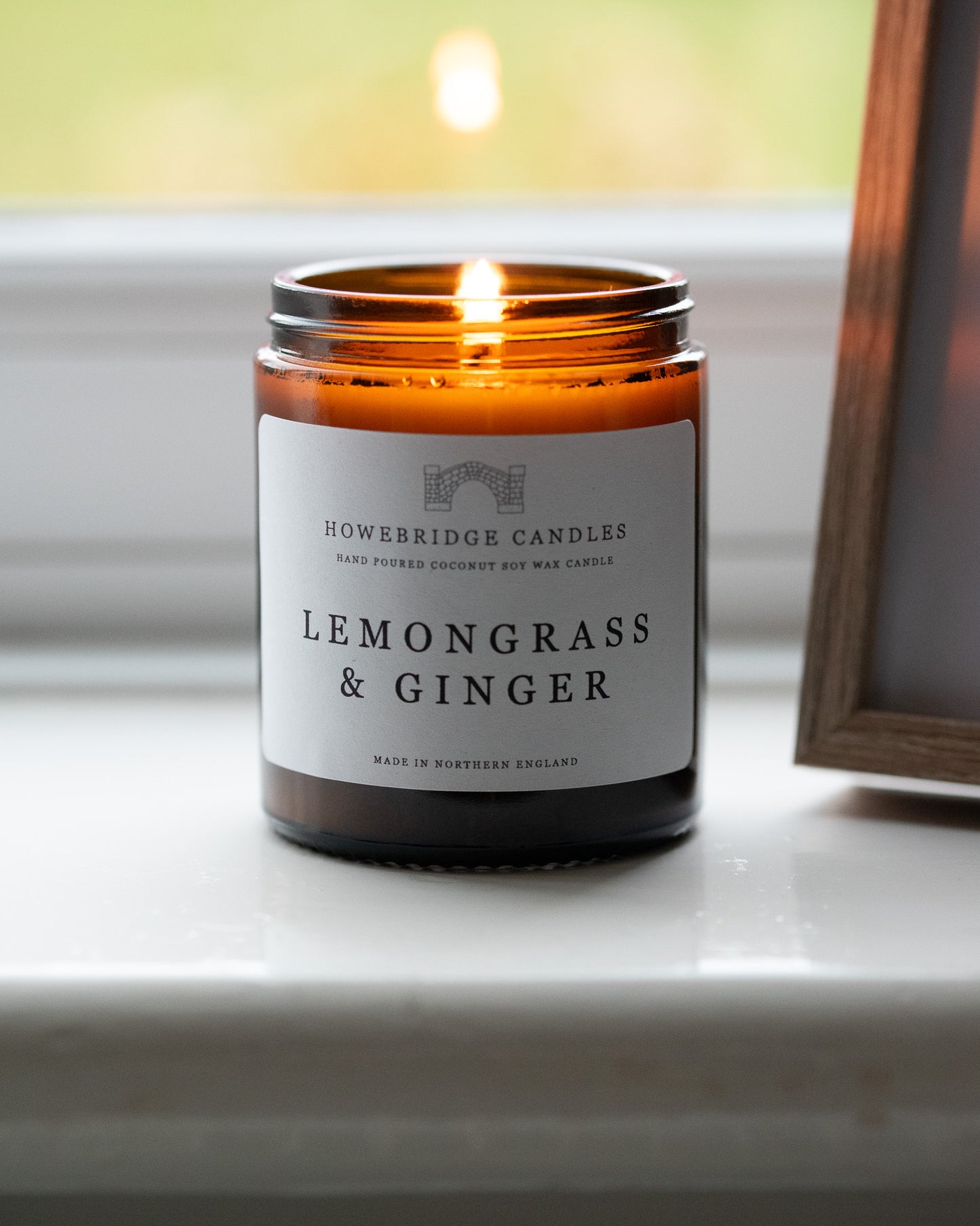Amber jar candle labeled 'Lemongrass & Ginger' by Howebridge Candles on a windowsill.