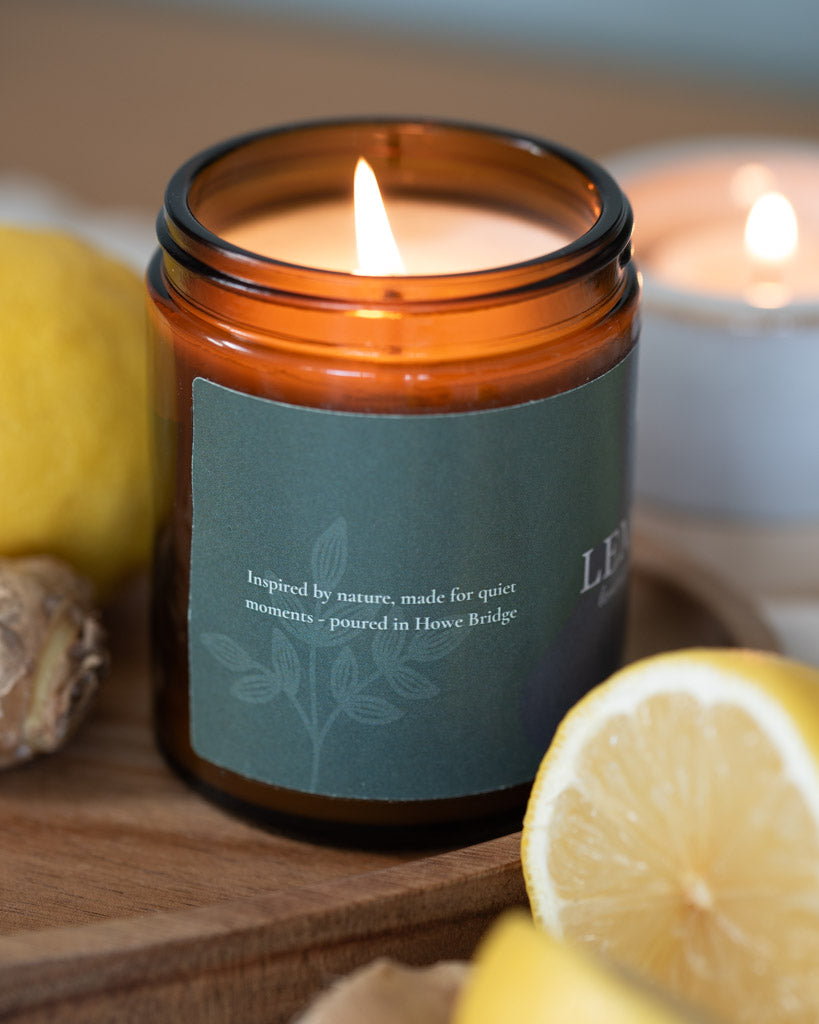 Lemon Peak candle lit surrounded by lemons and ginger. Sage green wrap around label, image shows side of label and says, "inspired by nature, made for quiet moments- poured in Howe Bridge'".