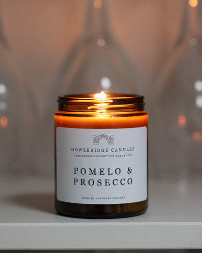 Pomelo & prosecco candle by Howebridge Candles. A lit, glass amber candle sat on a grey shelf.