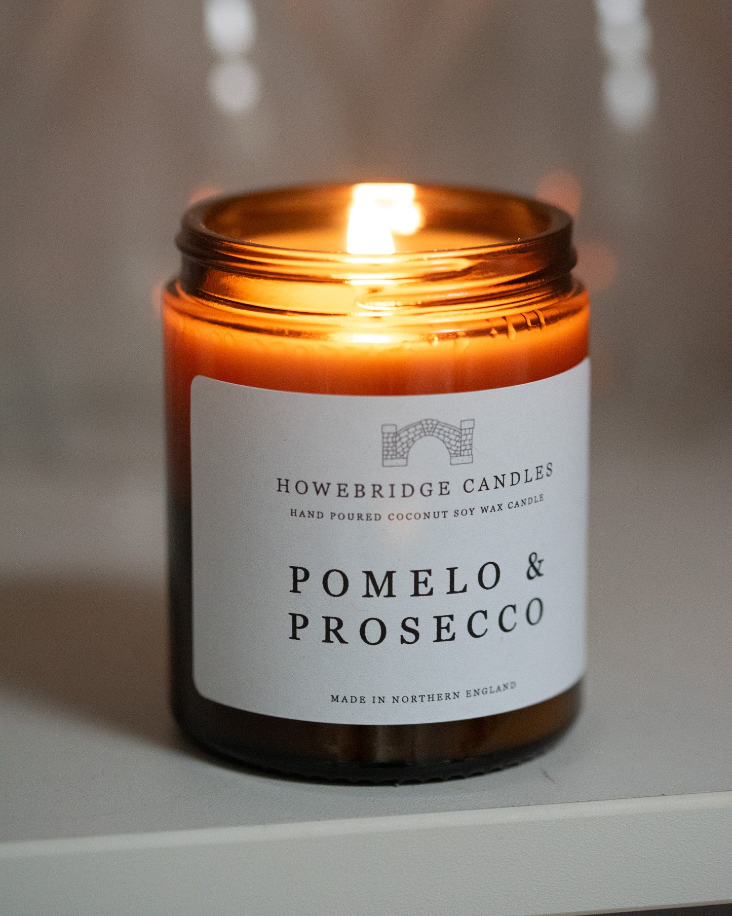 A amber glass candle by Howebridge Candles, with a white label and black writing with the scent pomelo & prosecco. 