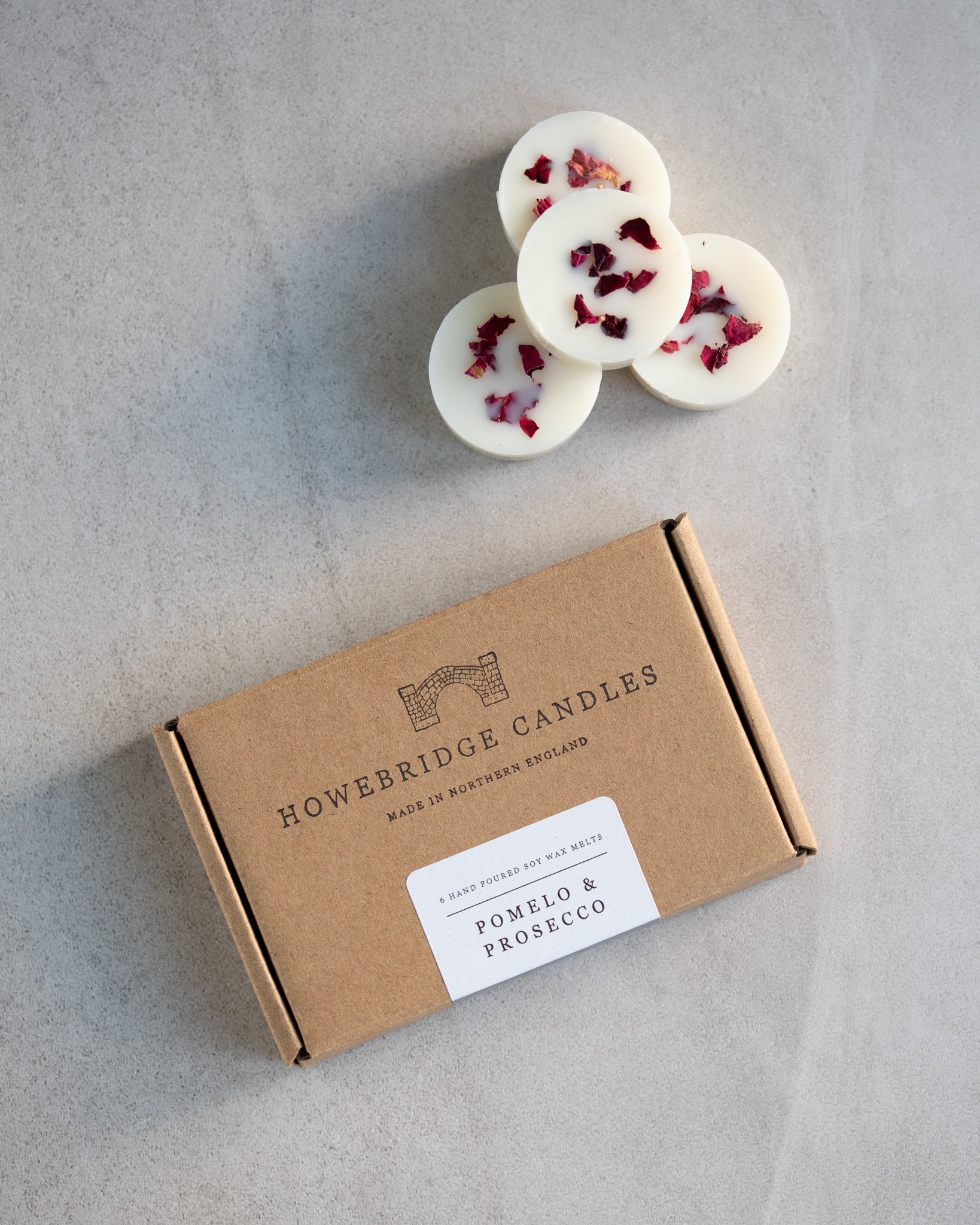Howebridge Candles pomelo & prosecco soy wax melts. Wax melts are stacked and sprinkled with dried rose petals. Next to them is the wax melt packaging, a kraft box stamped with their logo and sealed with a scent descriptive label.