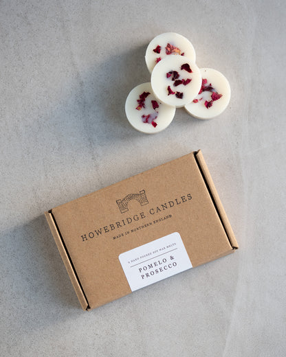 Howebridge Candles pomelo & prosecco soy wax melts. Wax melts are stacked and sprinkled with dried rose petals. Next to them is the wax melt packaging, a kraft box stamped with their logo and sealed with a scent descriptive label.