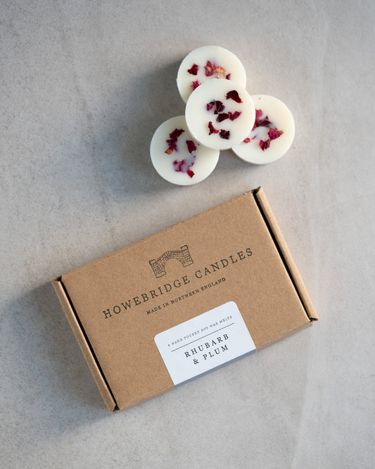Howebridge Candles rhubarb & plum soy wax melts. Showing a few stacked and sprinkled with dried rose petals. Next to them is the wax melts packaging, a kraft box stamped with their logo and sealed with a scent descriptive label.