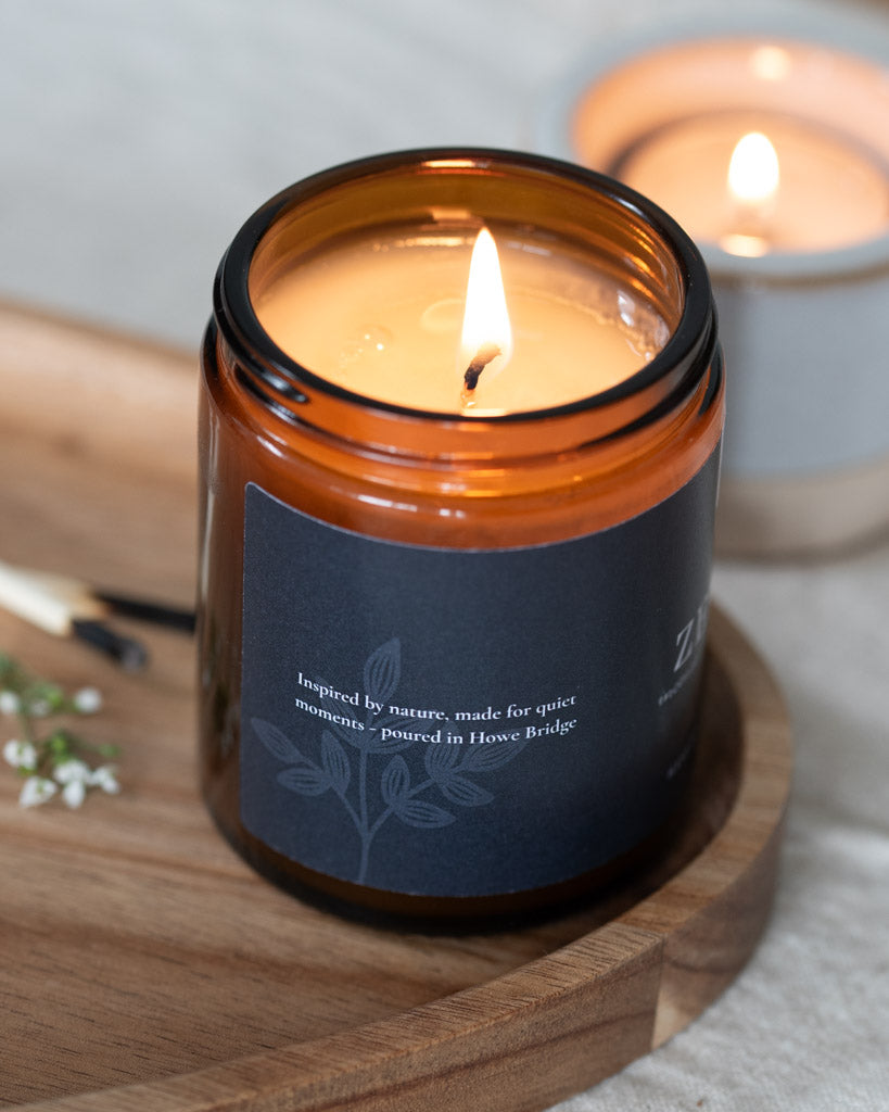 Candle in an amber glass jar with a wooden lid on a wooden surface. Botanical black candle label. Scent incense, sandalwood and frangipani. A rich Incense and slightly floral, the perfect candle to find inner balance, meditate or light during a yoga session.