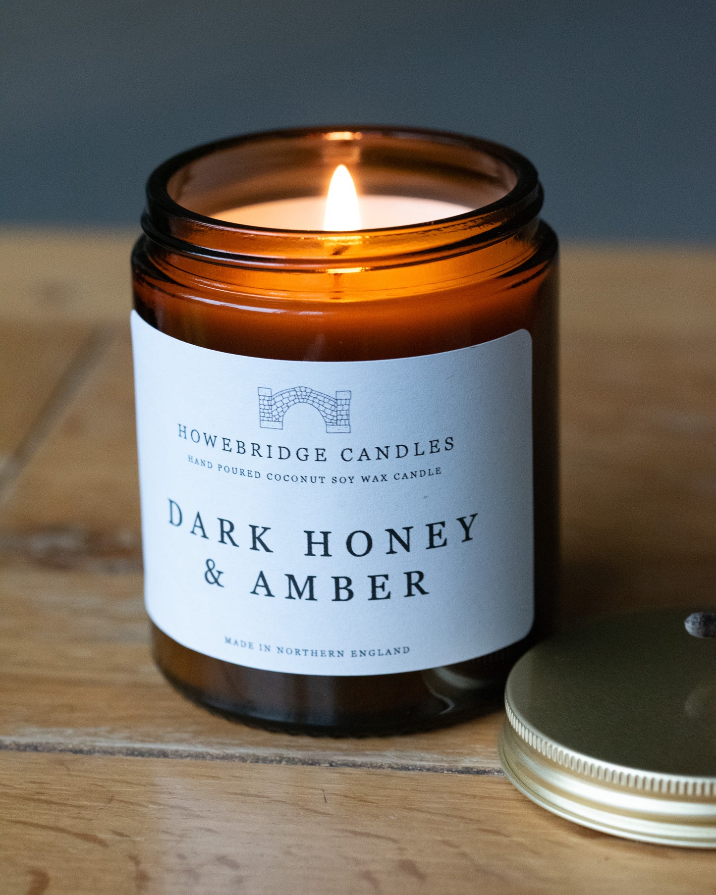 A amber glass candle called 'Dark honey & Amber' by Howebridge Candles, sat on a wooden table.