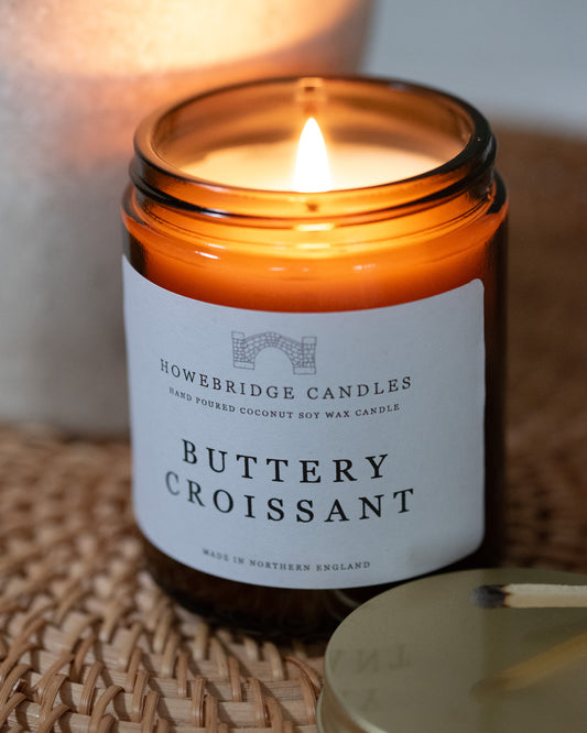 'Buttery Croissant' glass amber candle by Howebridge Candles, sat on a woven tray.