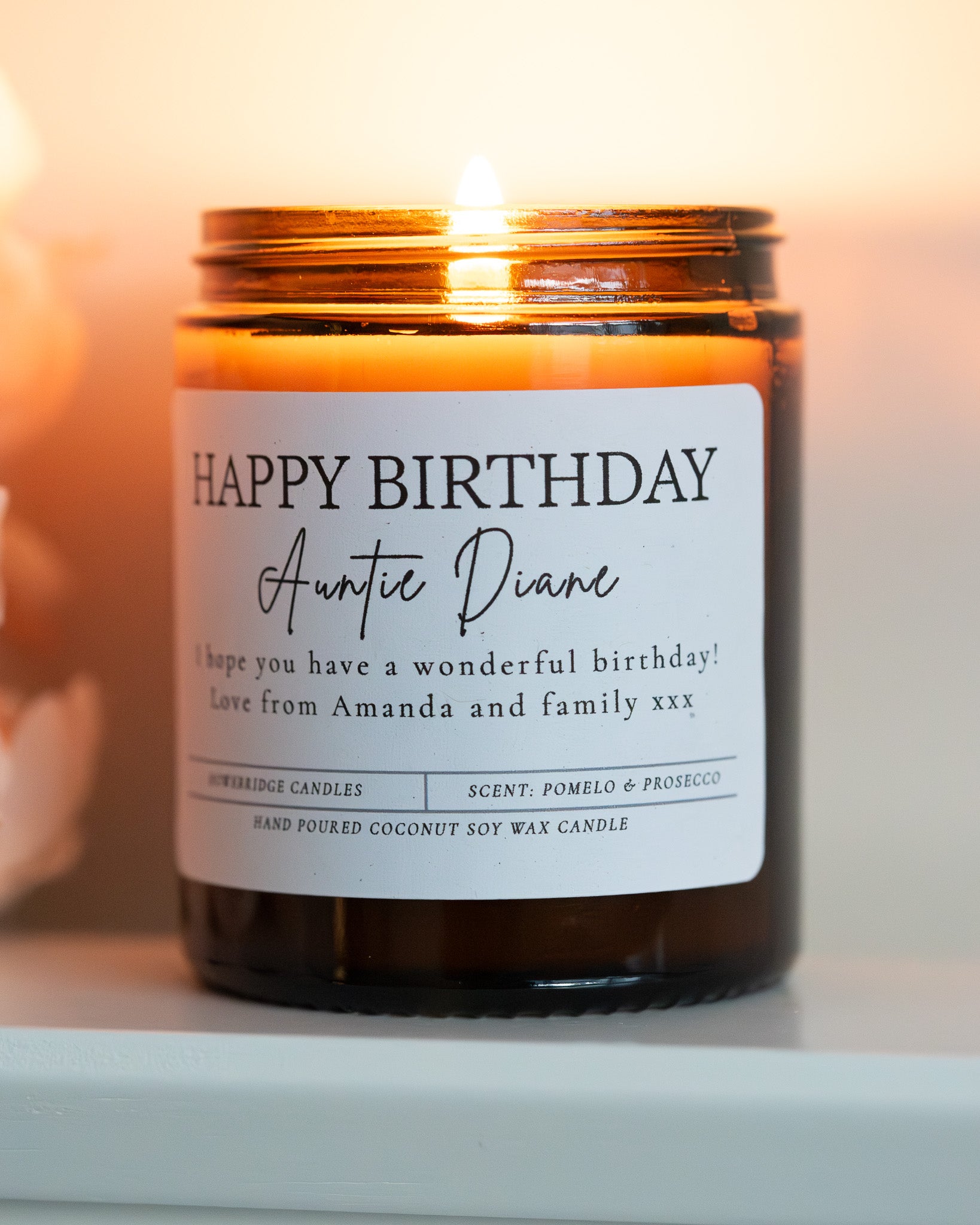 'Happy Birthday' Candle with name and message. Made by Howebridge Candles in a glass amber jar, it's lit and sat on a white shelf with pink flowers.