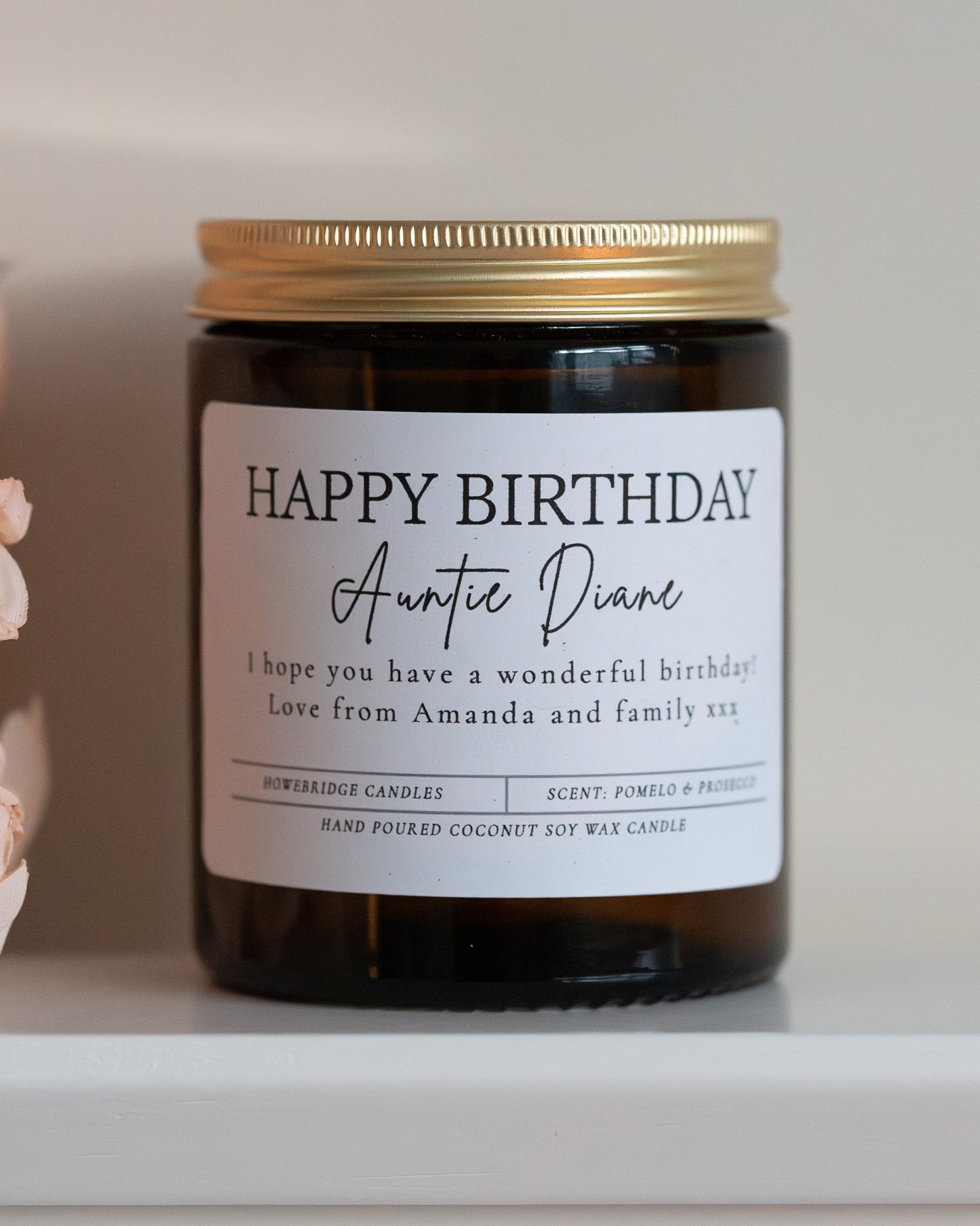 'Happy Birthday' Candle with name and personalised message. Made by Howebridge Candles in a glass amber jar and gold lid, sat on a white sheld with some pink flowers.