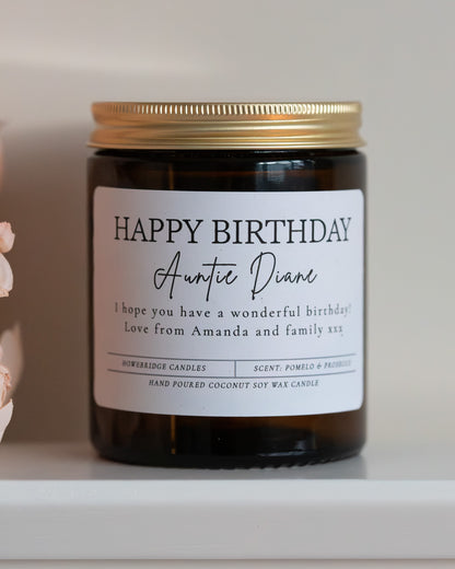 'Happy Birthday' Candle with name and personalised message. Made by Howebridge Candles in a glass amber jar and gold lid, sat on a white sheld with some pink flowers.
