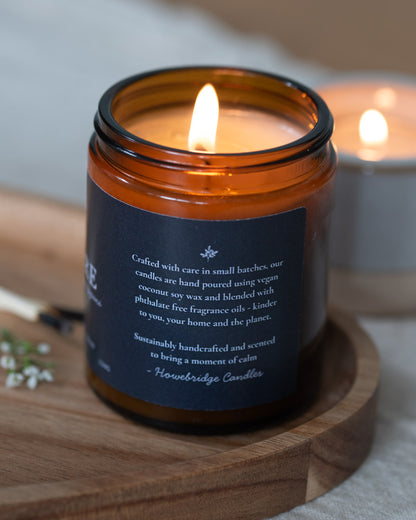 Candle in an amber glass jar with a wooden coaster on a light background. Black label with detail on the side. Scent incense, sandalwood and frangipani. A rich Incense and slightly floral, the perfect candle to find inner balance, meditate or light during a yoga session.