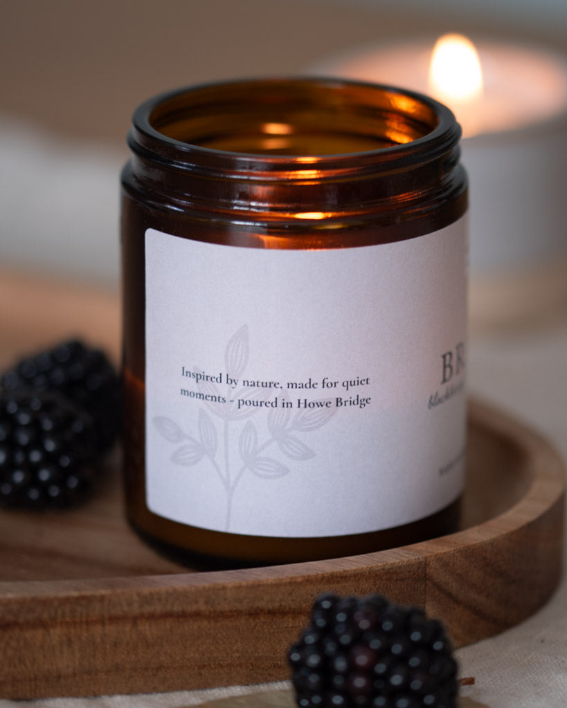 Candle in an amber jar with a white label on a wooden surface, surrounded by blackberries. Image is a close up of the side label and says "inspired by nature, made for quiet moments - poured in Howe Bridge.