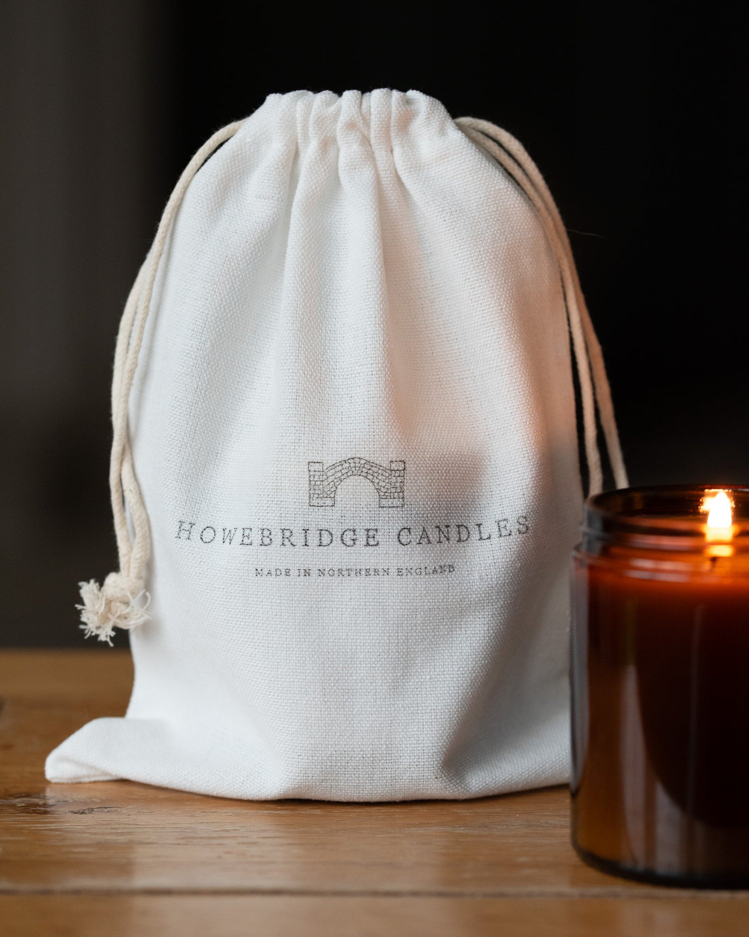 White drawstring bag with 'Howebridge Candles' branding next to a lit candle on a wooden surface.