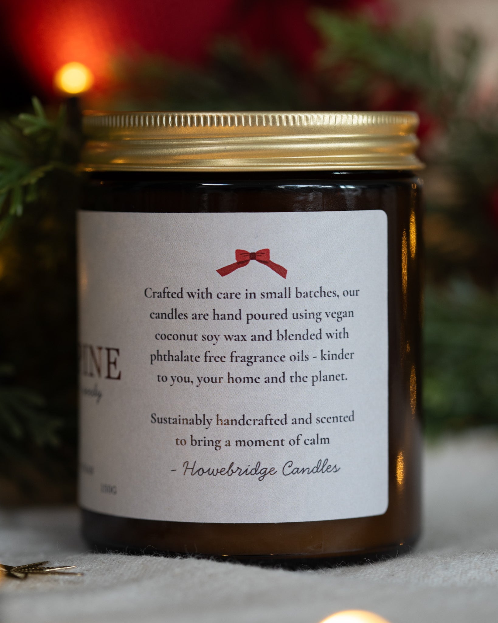A Christmas inspired wrap around candle label that features a red bow and information about the candle making process, such as being made in small batches, hand poured, vegan coconut soy wax and free from toxins. Poured in Howe Bridge, Atherton, UK.