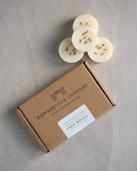 Howebridge Candles natural soy wax melts. Showing a few stacked and sprinkled with dry botanicals. Next to them is the wax melt packaging, stamped with their logo and sealed with a scent descriptive label.