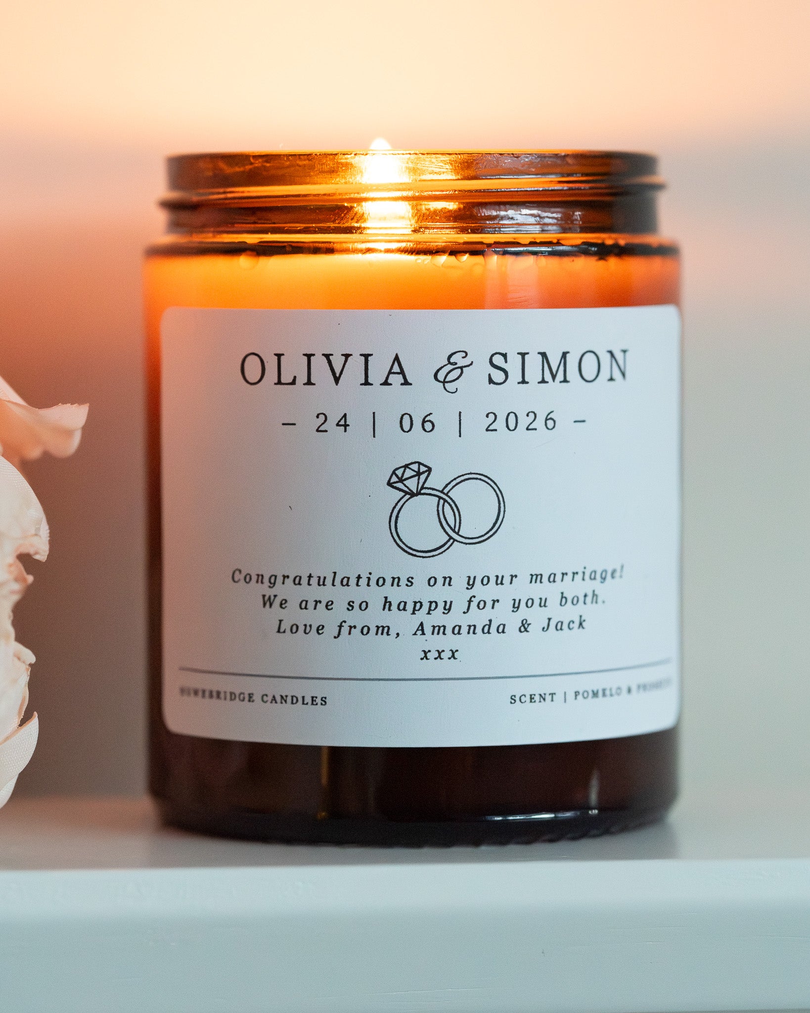 A personalised candle for a wedding day gift. Made by Howebridge Candles in a glass amber jar featuring a white label. Lit and sat on a white shelf with pink flowers.