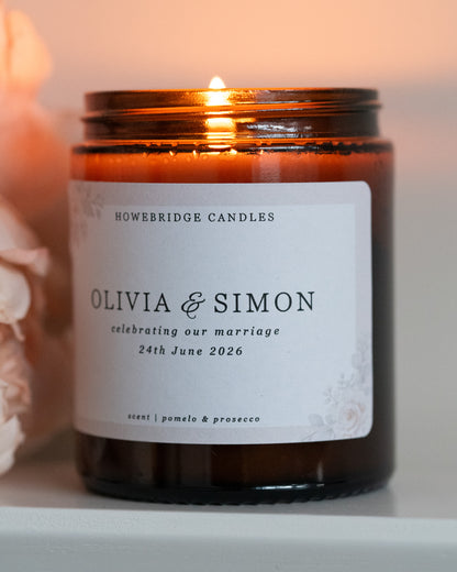 A wedding day candle by Howebridge Candles. A glass amber jar candle featuring a floral label. Lit and sat on a white shelf with pink flowers.