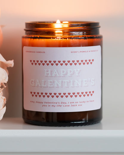 Glass Amber candle jar with pink 'Be My Valentine' label, lit and on a white surface, with some pink flowers.