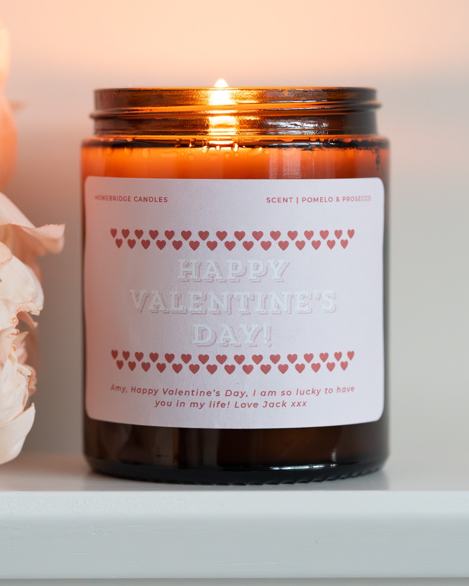 Glass Amber candle jar with pink 'Be My Valentine' label, lit and on a white surface, with some pink flowers.